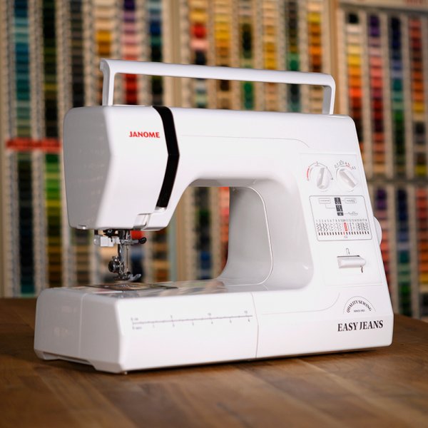 Janome Easy Jeans mekanisk symaskin står på ett bord framför ett skåp fullt av sytråd