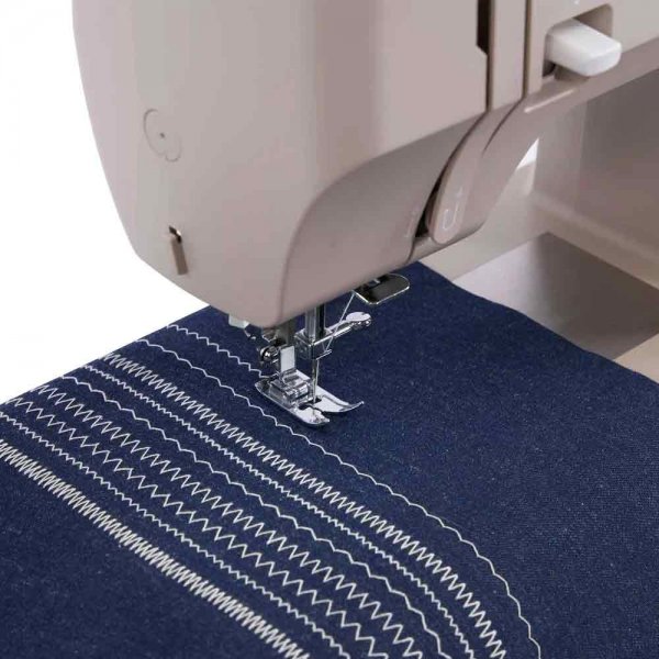 Singer symaskin i beige och sage, närbild på pressarfot och nål som syr vita sicksack- och mönstersömmar på mörkblått tyg.